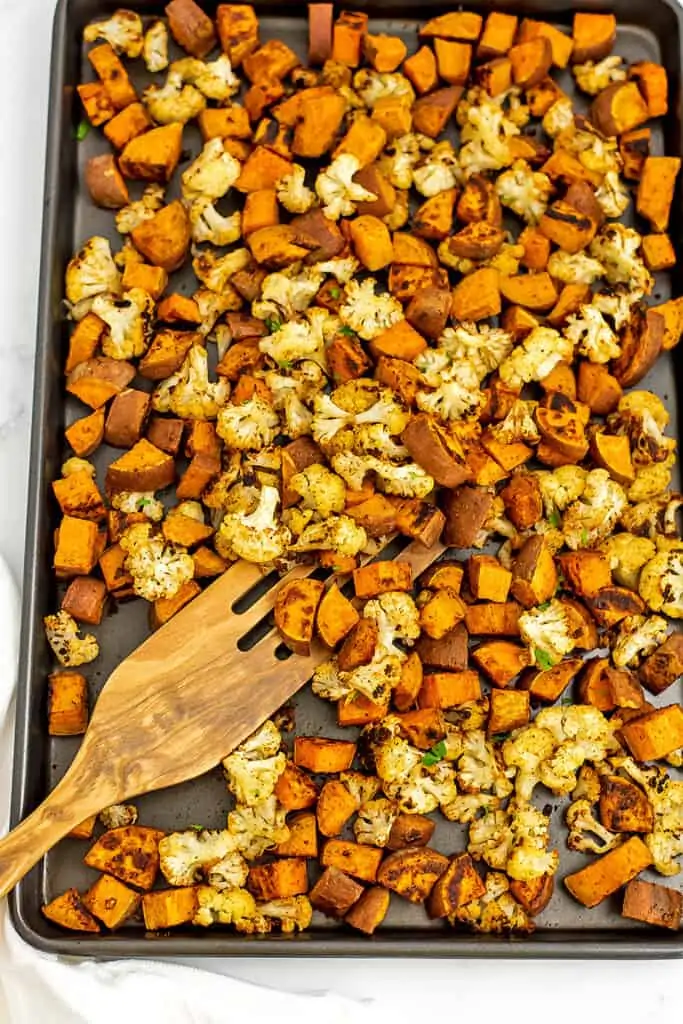 Sheet pan filled with roasted cauliflower and sweet potatoes with wooden spatula.