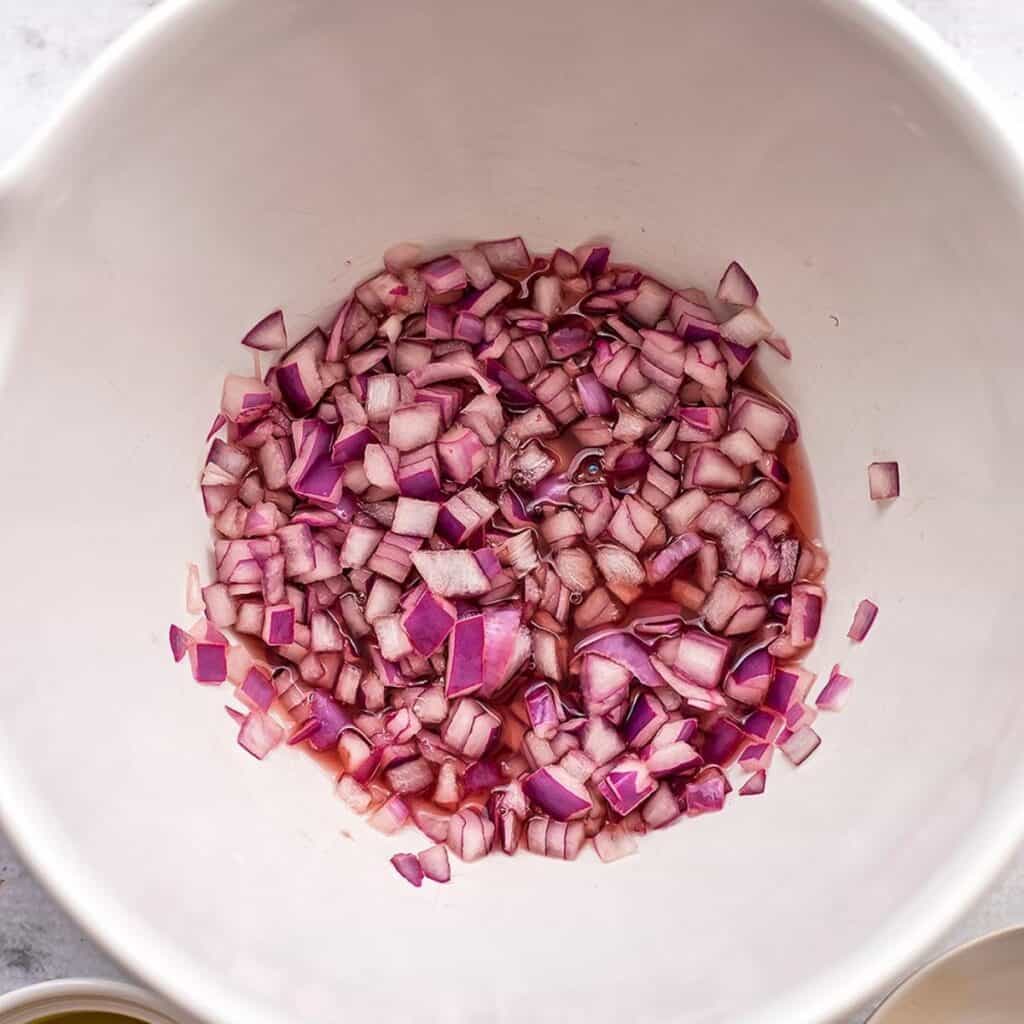 Red onions being soaked in red wine vinegar in white bowl.