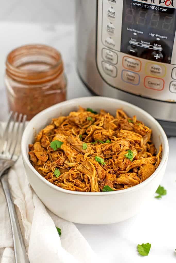 Shredded Mexican chicken in white bowl with instant pot in background.