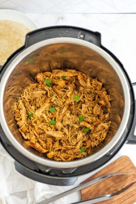 Taco shredded chicken in instant pot with tongs on wooden cutting board.