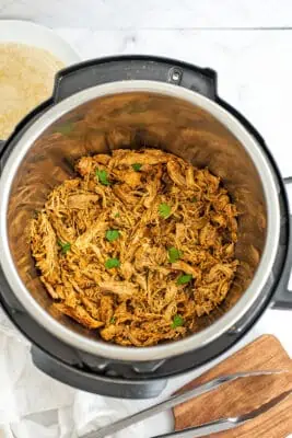 Taco shredded chicken in instant pot with tongs on wooden cutting board.