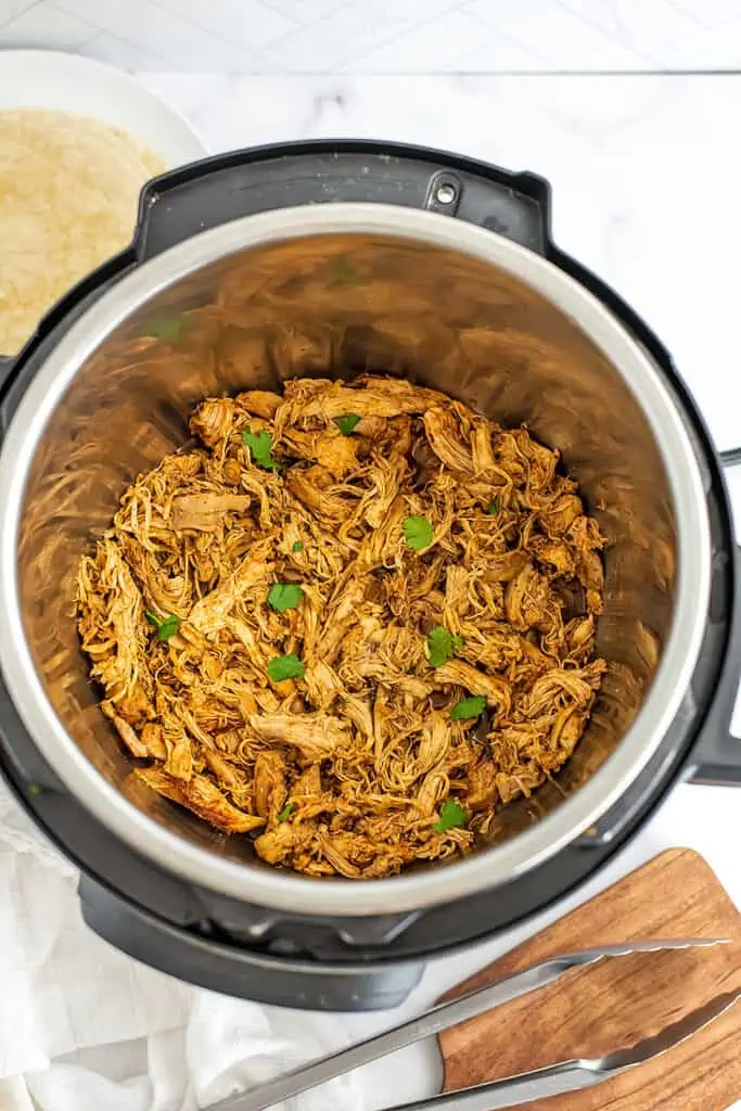 Taco shredded chicken in instant pot with tongs on wooden cutting board. 