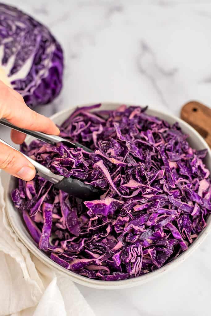 Red cabbage slaw, no mayo in white bowl, tongs being held in the slaw.