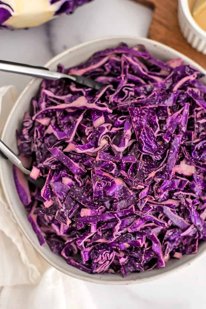 Tongs in a large white bowl filled with red cabbage slaw.