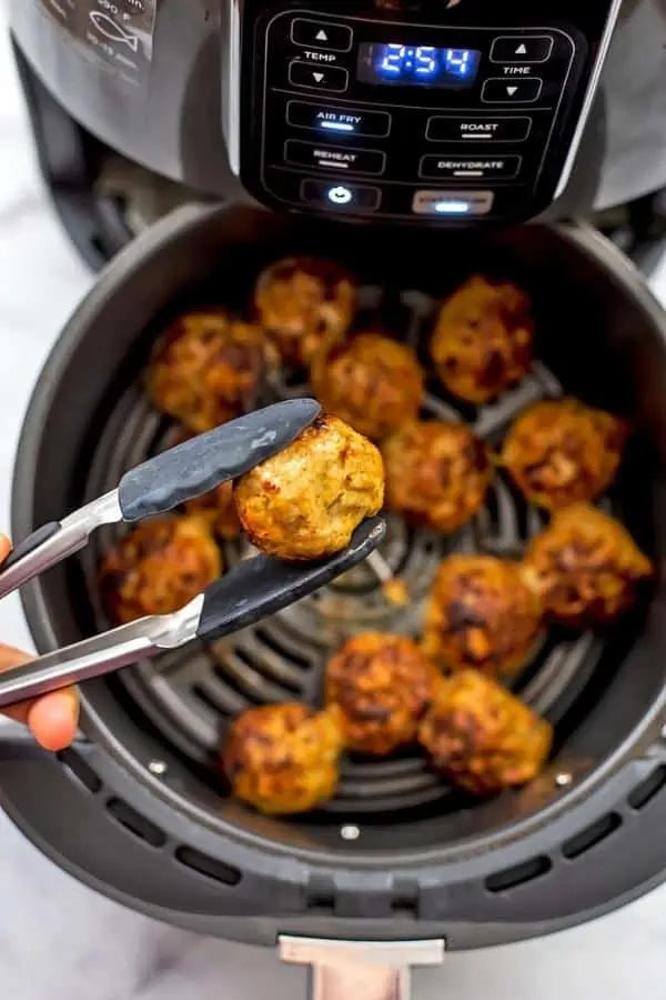 Tongs holding a chicken meatball over the air fryer basket.
