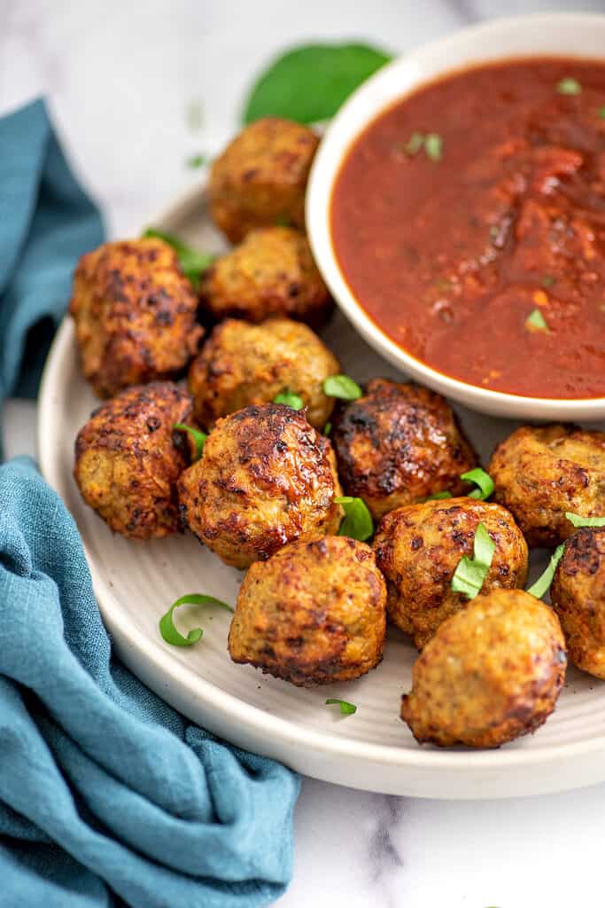 White plate filled with chicken meatballs cooked in the air fryer, bowl of marinara on the plate.