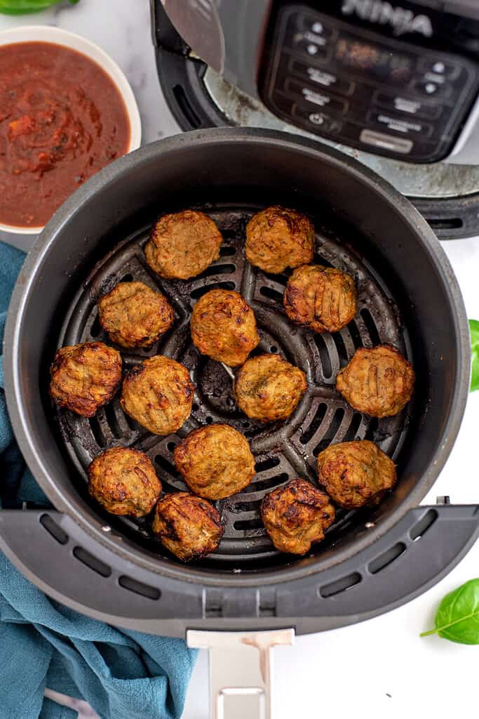 Air fryer basket filled with chicken meatballs, blue napkin and bowl of marinara on the side. 