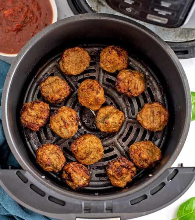 Air fryer basket filled with chicken meatballs, blue napkin and bowl of marinara on the side.