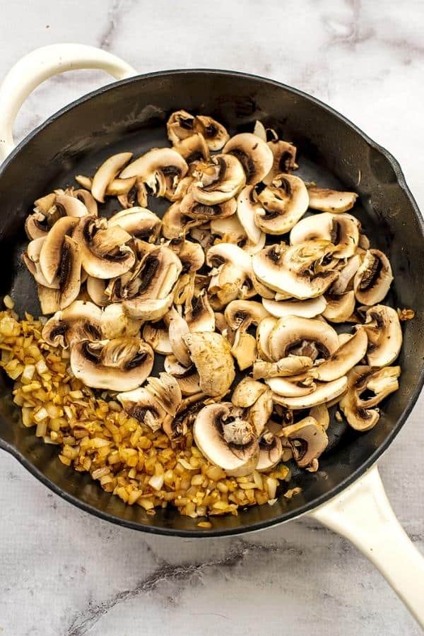Cast iron skillet with sliced mushrooms added, cooked onions pushed to the left side.