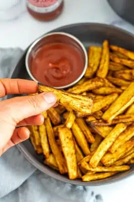Hand holding a parsnip fry over a plate full of fries.