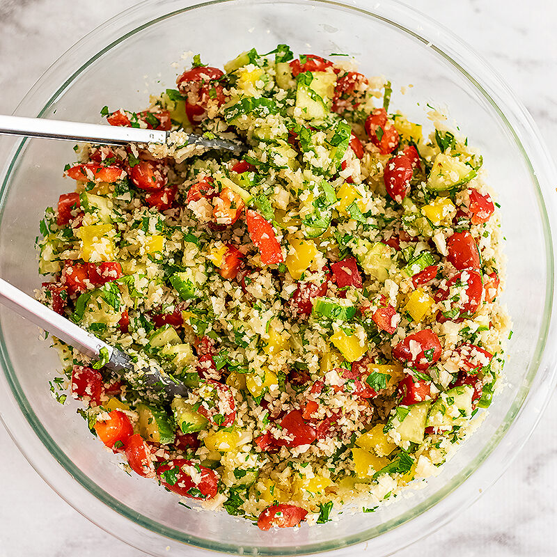 cauliflower tabbouleh in glass bowl after being mixed well. tongs resting in the bowl.