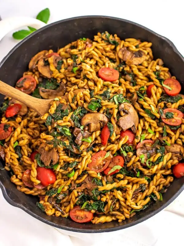 Wooden spoon in a skillet filled with balsamic kale mushroom pasta.