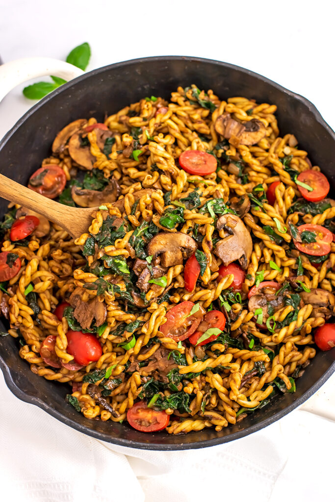 Wooden spoon in a skillet filled with balsamic kale mushroom pasta.