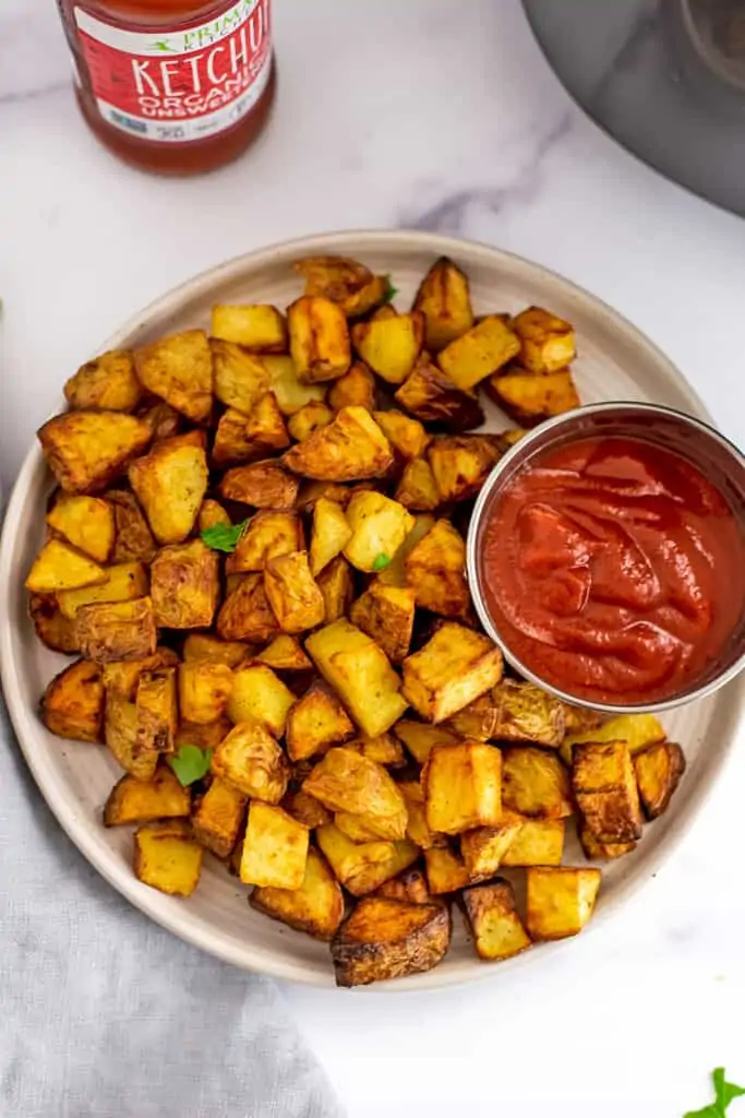 Air fryer diced potatoes on a white plate with a silver bowl of ketchup.