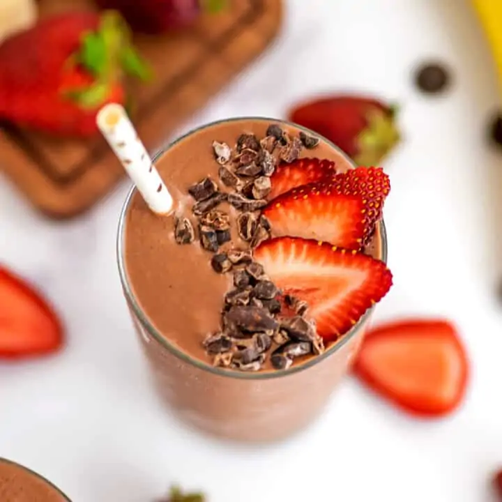 Strawberry banana chocolate smoothie in a glass with a straw, sliced strawberries and cacao nibs on top.