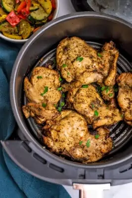 Greek chicken in an air fryer basket, veggies in the background.