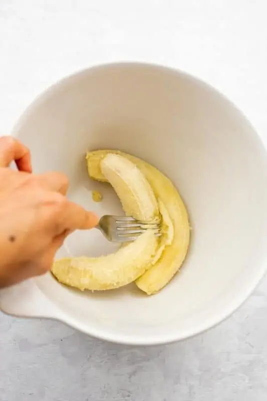 Hand holding a fork mashing bananas in large white bowl.