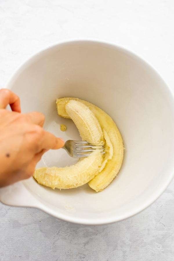 Hand holding a fork mashing bananas in large white bowl.