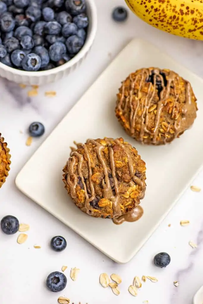 Banana blueberry oat muffins on white plate with almond butter drizzle on top.