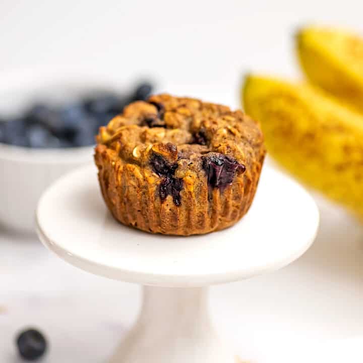 Banana blueberry oat muffin on a white muffin stand, bananas in background.