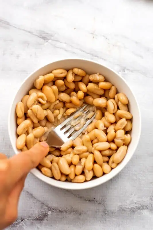 Fork mashing white beans in a white bowl.