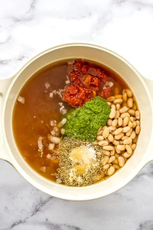 White pot filled with broth, white beans, pesto, diced tomatoes and spices.