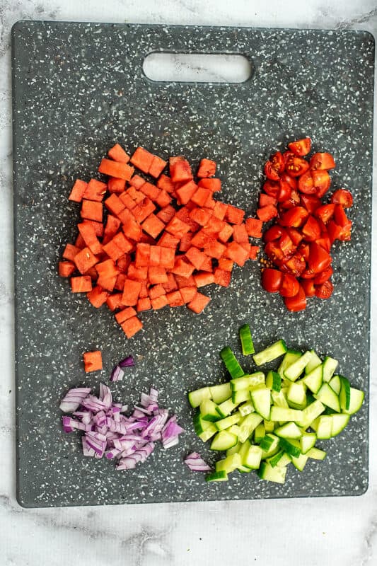 Watermelon, cucumber, red onion and tomatoes chopped into small pieces on cutting board.