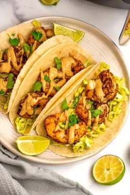 Three air fryer shrimp tacos on a white plate with limes.