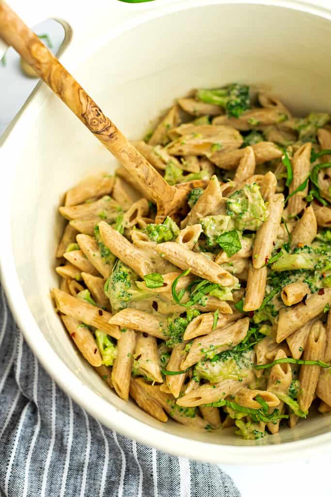 Wooden spoon in a white pot full of vegan broccoli pasta, blue striped napkin on the side.