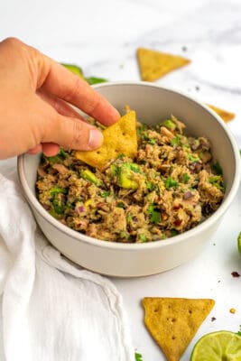 Hand dipping a chip into Mexican tuna salad in a white bowl.