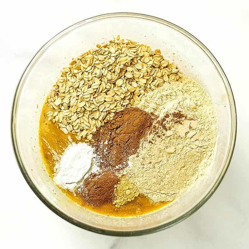 Rolled oats, spices, protein powder added to pumpkin mixture in glass bowl.