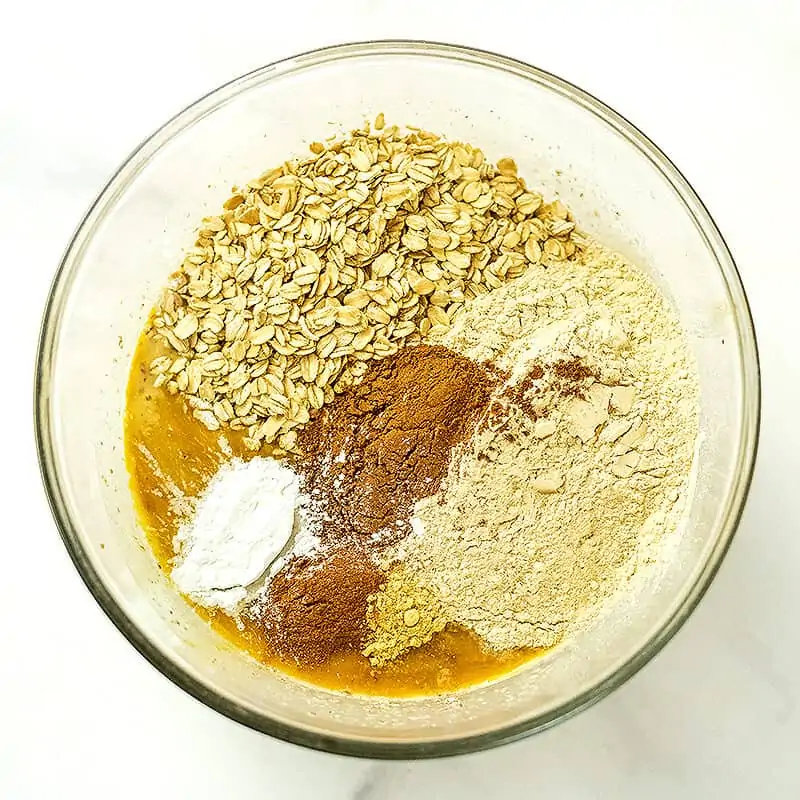 Rolled oats, spices, protein powder added to pumpkin mixture in glass bowl.