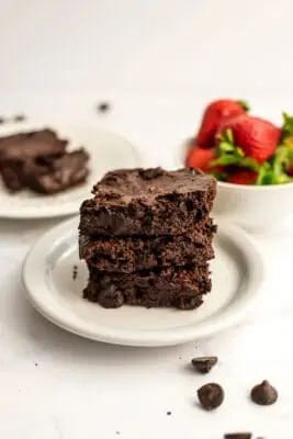 Three brownies made with oat flour on a white plate, strawberries in background.
