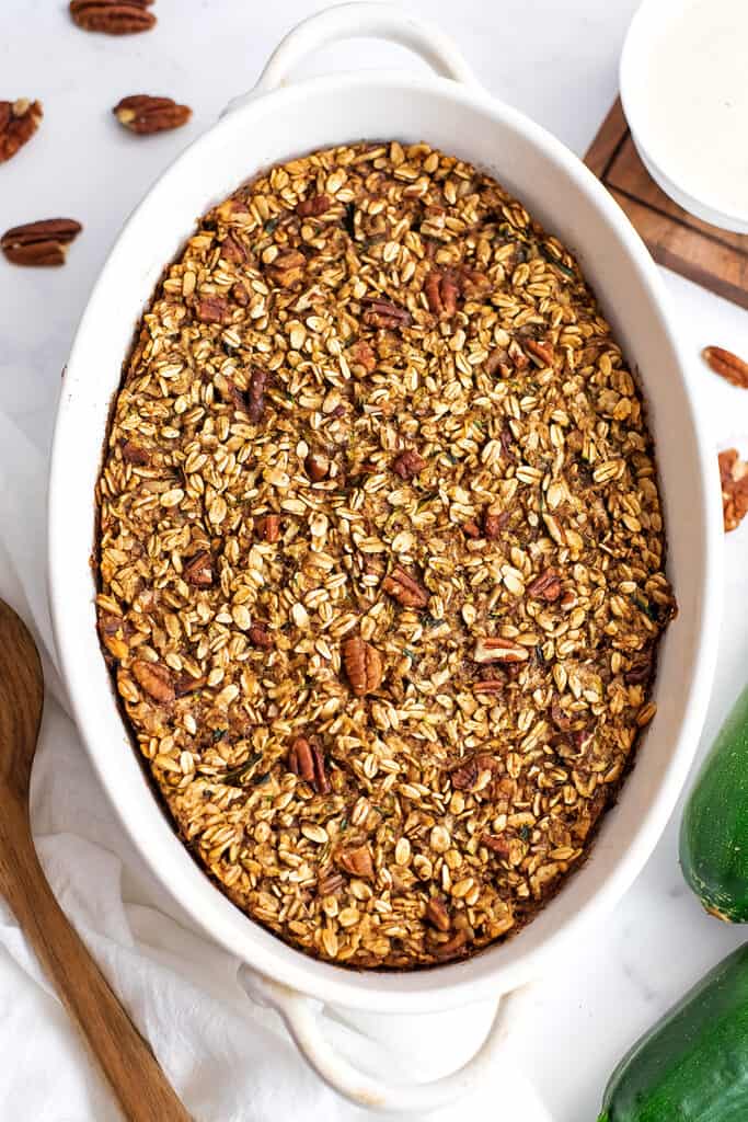 Zucchini baked oatmeal in a white casserole dish with pecans and zucchini in the background.