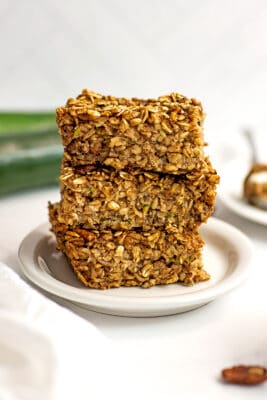 Three slices of zucchini baked oatmeal stacked on a white plate, zucchini in background.