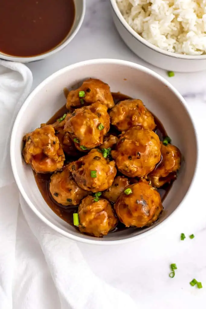 Chicken meatballs covered in sweet and sour sauce in a white bowl with sweet and sour sauce behind.