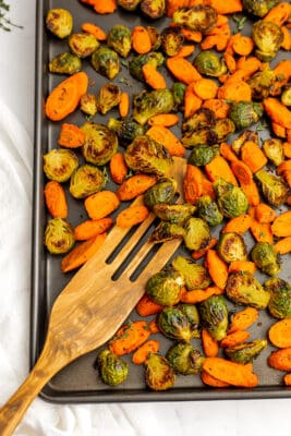 Baking sheet filled with roasted carrots and brussel sprouts and a wooden spatula.