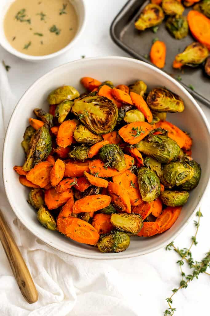 Roasted carrots and brussel sprouts in a white bowl, with tahini sauce in the background.