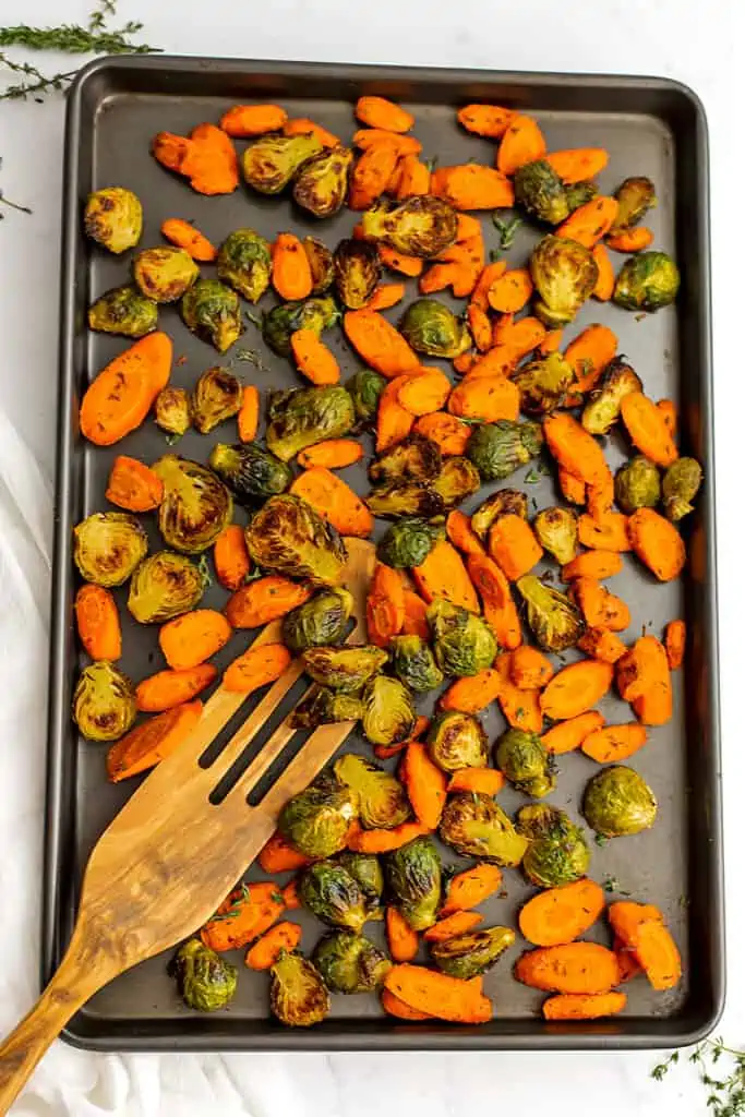 Baking sheet filled with roasted brussel sprouts with carrots with wooden spatula on the side.