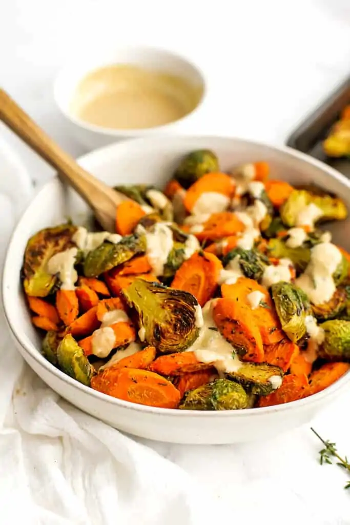 Roasted brussel sprouts and carrots in a white bowl with tahini drizzled on top.