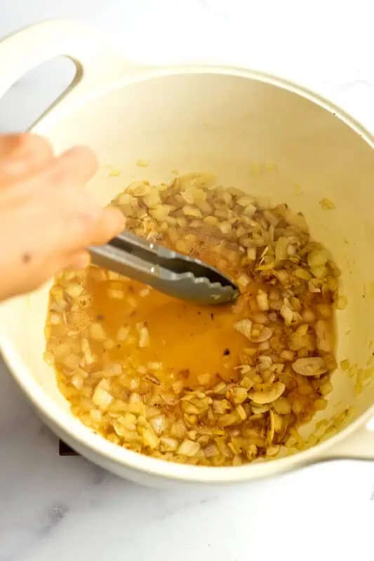 Tongs moving around the onions and garlic in pot with some vegetable broth added.