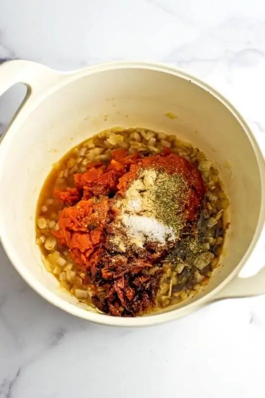 Tomatoes, sundried tomatoes and spices added to cooked onions in white pot.