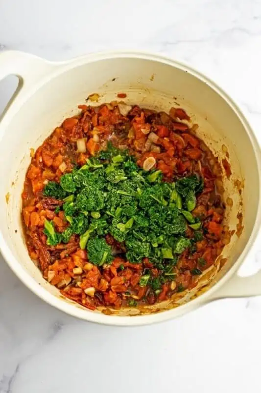 Frozen kale being added to tomato mixture in large white pot.