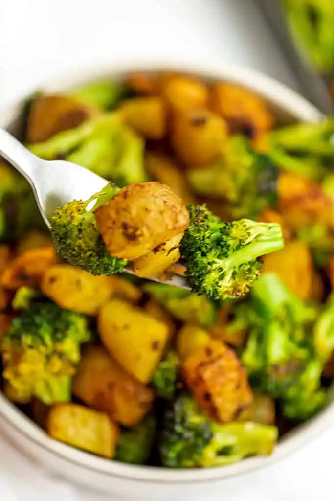 Fork holding roasted broccoli and potatoes over a white bowl. 