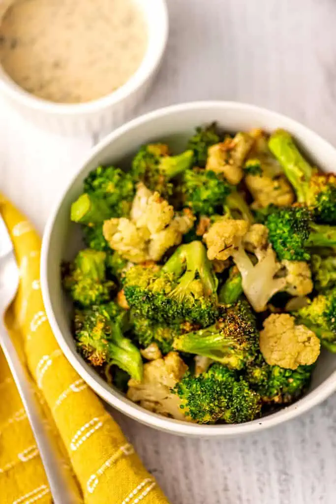 Air fryer cauliflower and broccoli in a white bowl with tahini dip in the background.