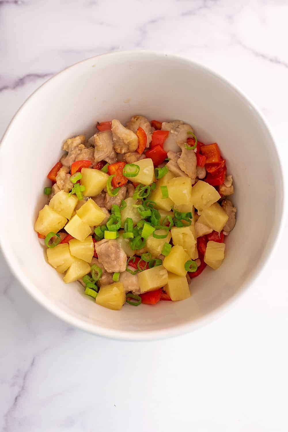 Pineapple chunks added to diced chicken and chopped bell peppers in white bowl.