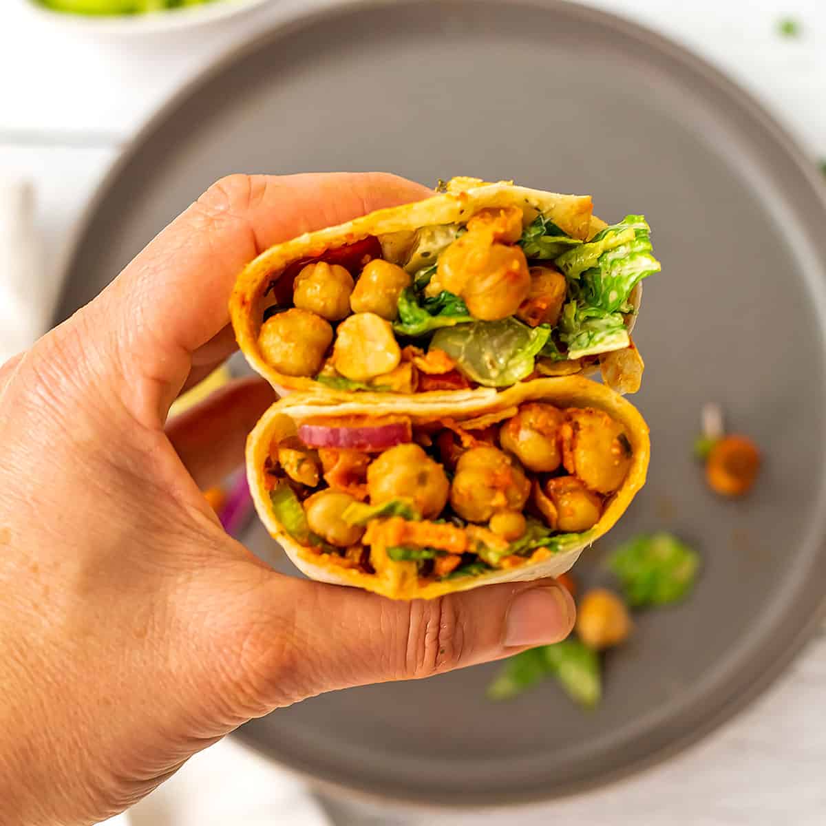 Buffalo chickpea wrap cut in half being held by a hand over grey plate.