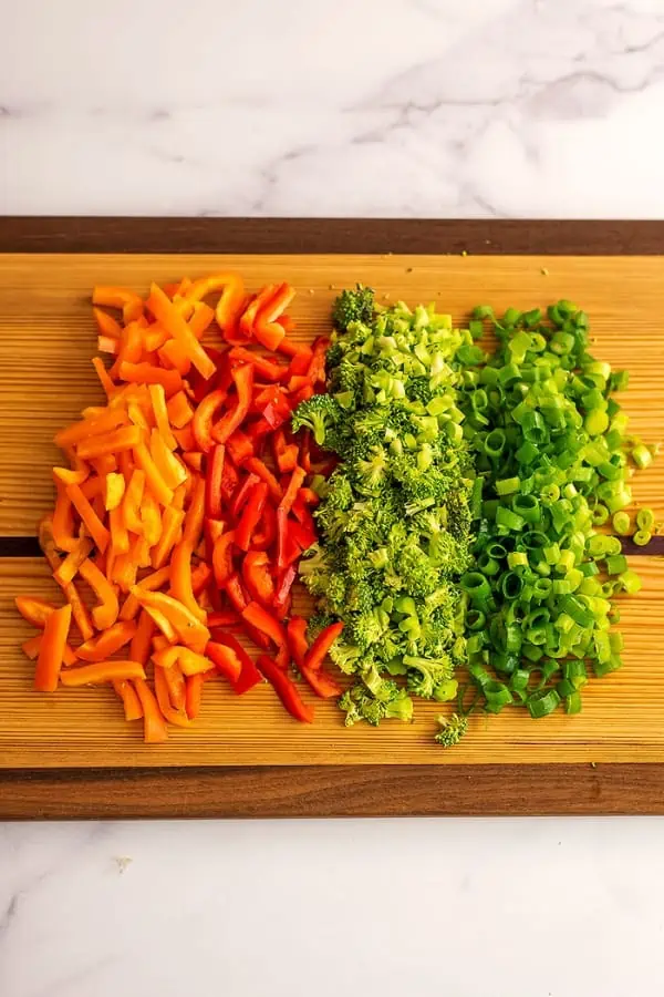 Sliced bell peppers, chopped broccoli and green onions on wood cutting board.