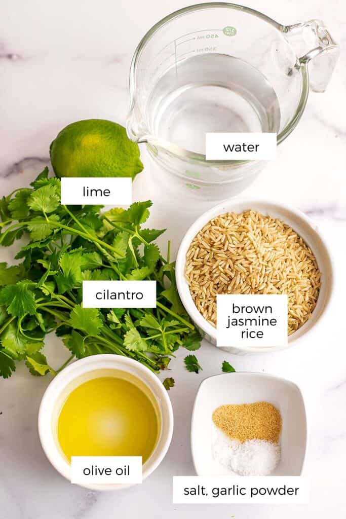 Ingredients to make cilantro lime brown rice in white bowls on marble countertop.