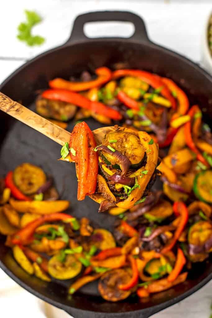 Wooden spoonful of fajita vegetables over cast iron skillet.
